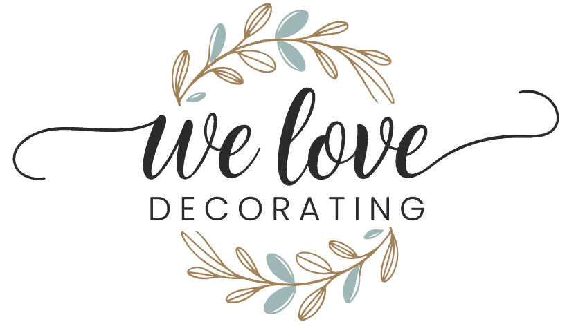 A decorative logo. At the center, there's a wreath made of leaves in shades of blue and gold. Surrounding the wreath is a cursive script that reads 'we love decorating'. The text is in black, and the wreath and the text are positioned centrally, making them the focal points of the design.